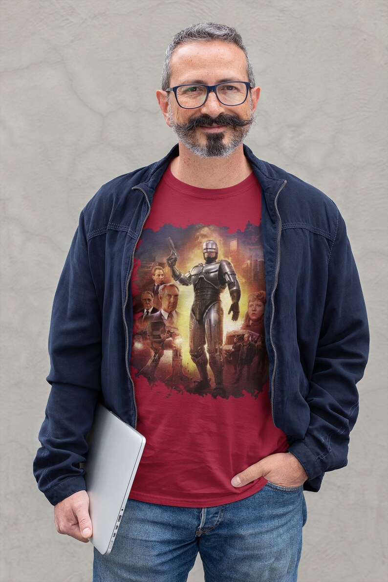 May include: A man wearing a red t-shirt with a graphic of Robocop and other characters from the movie. He is also wearing a blue jacket and jeans. He is holding a laptop computer in his left hand.