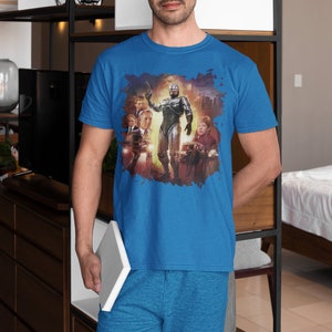 May include: A blue t-shirt with a graphic print of a robot and other characters from the movie Robocop. The shirt is being worn by a person standing in front of a bookshelf.
