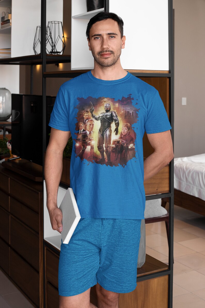 May include: A blue t-shirt with a graphic print of a robot and other characters from the movie Robocop. The shirt is being worn by a person standing in front of a bookshelf.