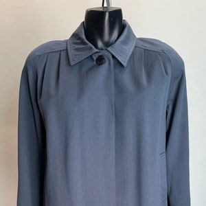 Puede incluir: Un abrigo largo azul pizarra con un diseño clásico. El abrigo presenta un cuello con solapas, un cierre de un solo botón negro y mangas largas. La tela tiene una sutil textura vertical, y el abrigo se exhibe en un maniquí.