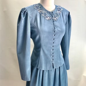 Może przedstawiać: Jas i spódnica w jasnoniebieskim kolorze z ozdobnym dekoltem z koralików. Kurtka zapinana na guziki z przodu i długie rękawy. Spódnica o kroju A z plisowanym wzorem.