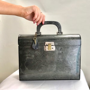 Könnte beinhalten: Ein vintage grauer Metallkoffer mit einem schwarzen Griff und einem silbernen Schloss. Der Koffer hat eine strukturierte Oberfläche und einen daran befestigten Schlüssel.