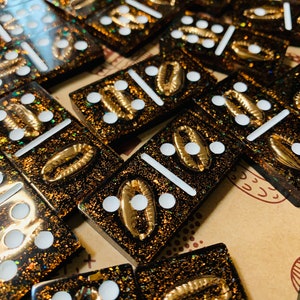 May include: A set of black dominoes with gold glitter and white dots. Each domino has a gold seashell design on one side.