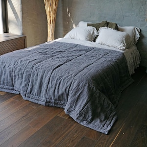 May include: A bed with a grey linen quilt and white pillows. The bed is on a dark wood floor. A tall bundle of dried plants is in the background, next to a window.