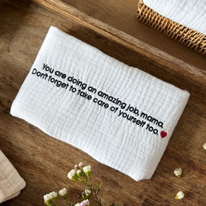 May include: A white cotton dish towel with the embroidered text "You are doing an amazing job, mama. Don't forget to take care of yourself too." and a small red heart.