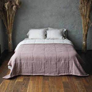 May include: A bed with a dusty rose quilt and white pillows. The bed has a light grey sheet. The room has a dark wood floor and a grey wall. Tall dried plants are in vases either side of the bed.