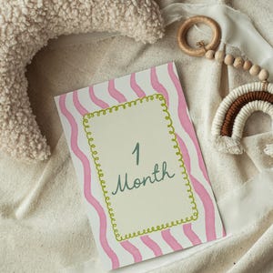 May include: A baby milestone card with pink wavy stripes and a green border, displaying the text "1 Month" in a handwritten style. A plush crescent moon pillow, a wooden ring with beads, and a macrame rainbow are also in the image.