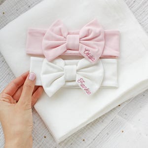 Pode incluir: Duas faixas de tecido macio com laços, uma em rosa claro e outra em branco. A faixa rosa apresenta o nome bordado Odilia, e a faixa branca apresenta o nome bordado Frankie numa fonte cursiva rosa a condizer.