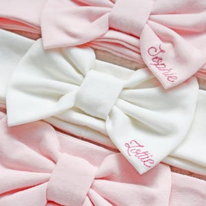 Op de afbeelding: Close-up van verschillende baby haarbanden in zachtroze en wit. Elke haarband heeft een grote strik. Twee roze haarbanden zijn geborduurd met de namen "Sophie" en "Lottie" in roze draad. De haarbanden zijn gemaakt van een zachte, rekbare stof.
