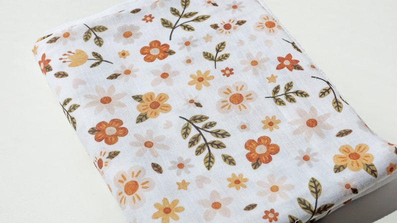 Op de afbeelding: Witte stof met een bloemenpatroon met oranje, gele en groene bloemen. De stof is dubbelgevouwen.