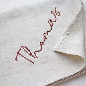 May include: A folded, light beige linen cloth with the name "Thomas" embroidered in a cursive, reddish-brown thread. The fabric has a natural, textured appearance, and the stitching is neat and detailed.