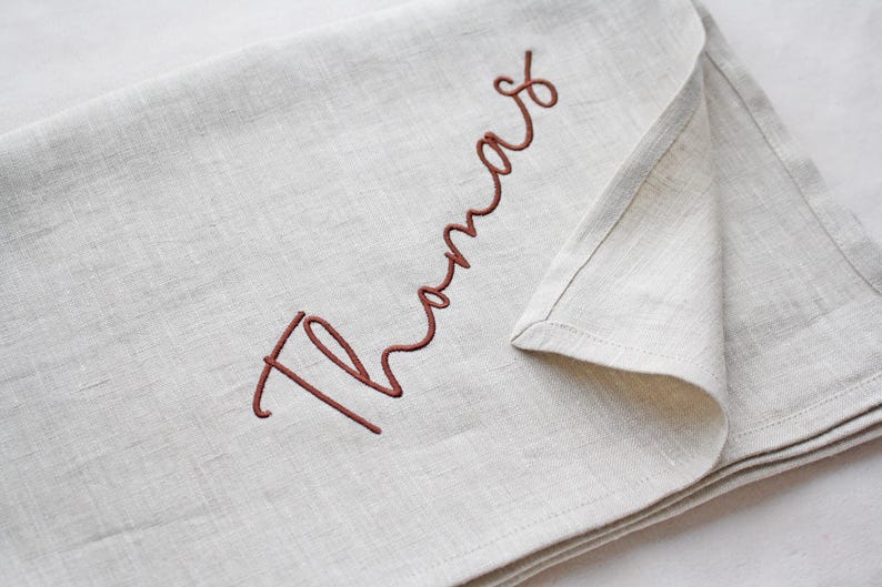 May include: A folded, light beige linen cloth with the name "Thomas" embroidered in a cursive, reddish-brown thread. The fabric has a natural, textured appearance, and the stitching is neat and detailed.