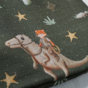 Op de afbeelding: Groene stof met een herhalend patroon van een vos die op een dinosaurus rijdt, sterren en palmbomen.