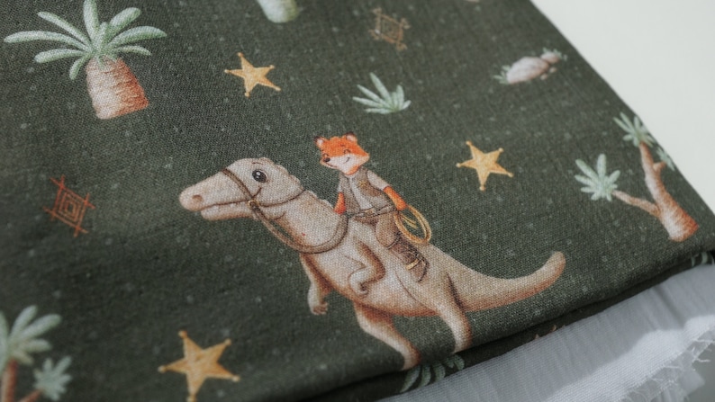 Op de afbeelding: Groene stof met een herhalend patroon van een vos die op een dinosaurus rijdt, sterren en palmbomen.