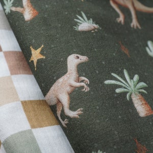 Op de afbeelding: Een groene stof met een herhalend patroon van dinosaurussen, sterren en cactussen. De stof is gevouwen over een bruin en wit geruite stof.