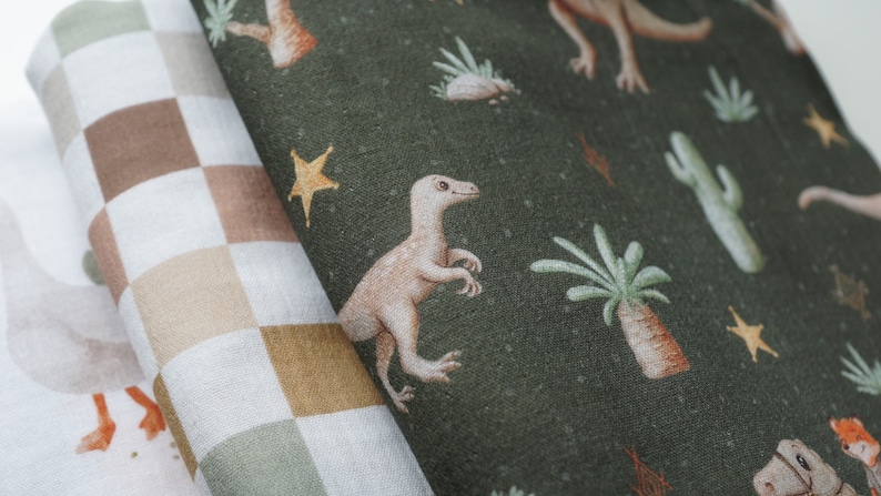 Op de afbeelding: Een groene stof met een herhalend patroon van dinosaurussen, sterren en cactussen. De stof is gevouwen over een bruin en wit geruite stof.