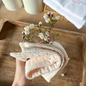 May include: A soft, peach-colored, folded cloth towel with a textured surface. The towel is held in a hand and is surrounded by small white flowers.