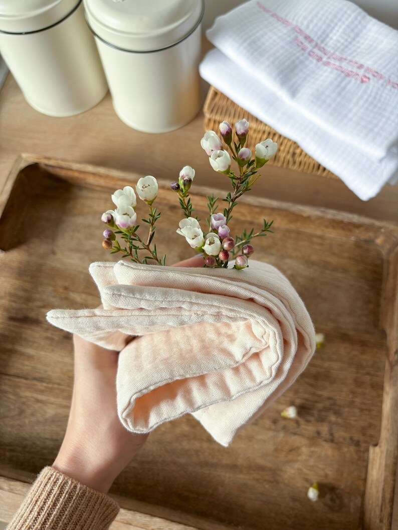 May include: A soft, peach-colored, folded cloth towel with a textured surface. The towel is held in a hand and is surrounded by small white flowers.