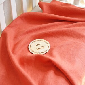 May include: Coral-colored linen baby blanket draped over a crib mattress. A wooden disc with the text "One Month" is centered on the blanket. The blanket is soft and appears to be made of natural fibers.