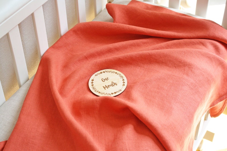May include: Coral-colored linen baby blanket draped over a crib mattress. A wooden disc with the text "One Month" is centered on the blanket. The blanket is soft and appears to be made of natural fibers.