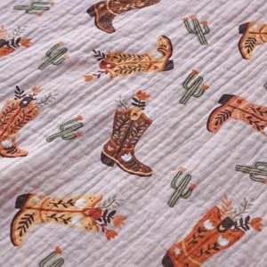 May include: A pink fabric with a repeating pattern of brown cowboy boots with floral accents and green cactus plants.