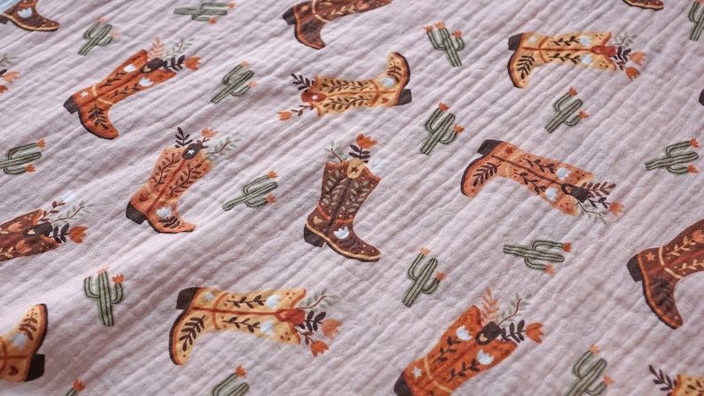 May include: A pink fabric with a repeating pattern of brown cowboy boots with floral accents and green cactus plants.