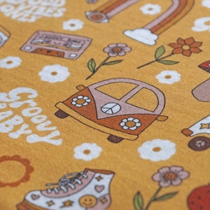 May include: A yellow fabric with a groovy baby print. The print features a variety of 1970s inspired designs, including a peace sign, a van, a mushroom, a cassette tape, a sun, and flowers. The text "Groovy Baby" is written in a retro font.