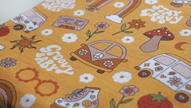 May include: A yellow fabric with a groovy baby print. The print features a variety of 1970s inspired designs, including a peace sign, a van, a mushroom, a cassette tape, a sun, and flowers. The text "Groovy Baby" is written in a retro font.