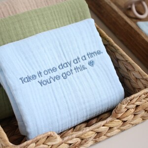 May include: A blue muslin baby blanket with the embroidered text "Take it one day at a time. You've got this."  The blanket is folded and placed in a woven basket.