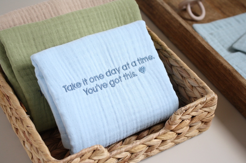 May include: A blue muslin baby blanket with the embroidered text "Take it one day at a time. You've got this."  The blanket is folded and placed in a woven basket.