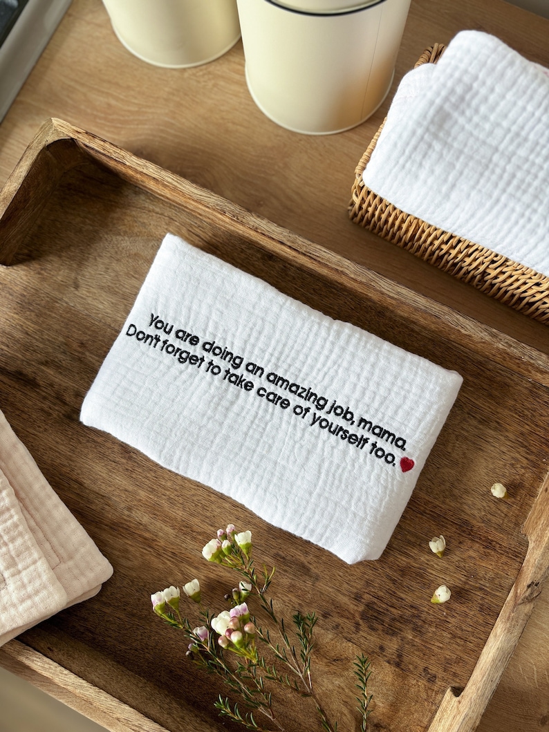 May include: A white cotton dish towel with the embroidered text "You are doing an amazing job, mama. Don't forget to take care of yourself too." and a small red heart.