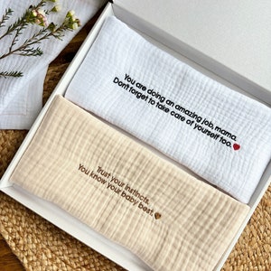 May include: Two white muslin baby burp cloths with embroidered messages. The top cloth reads "You are doing an amazing job, mama. Don't forget to take care of yourself too." The bottom cloth reads "Trust your instincts. You know your baby best."