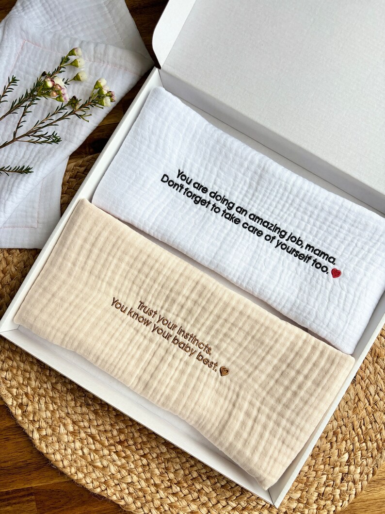 May include: Two white muslin baby burp cloths with embroidered messages. The top cloth reads "You are doing an amazing job, mama. Don't forget to take care of yourself too." The bottom cloth reads "Trust your instincts. You know your baby best."
