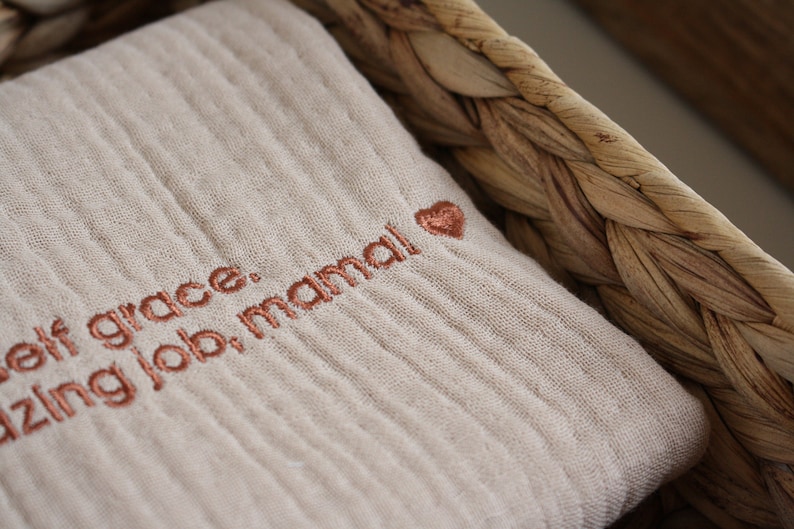 May include: A beige muslin fabric square with the text "Self grace. Amazing job, mama!" embroidered in copper-colored thread with a small heart.