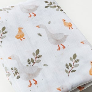 Op de afbeelding: Een witte muslin swaddle deken met een herhalend patroon van grijze ganzen en oranje eendjes met groene bladeren.