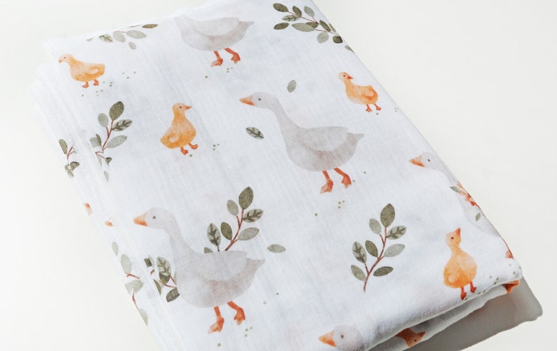 Op de afbeelding: Een witte muslin swaddle deken met een herhalend patroon van grijze ganzen en oranje eendjes met groene bladeren.