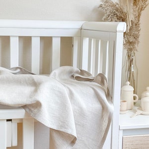May include: A white crib with a light beige linen blanket draped over the side. The blanket has a soft, textured appearance. A vase of dried pampas grass and a small tea set are on a nearby white table.