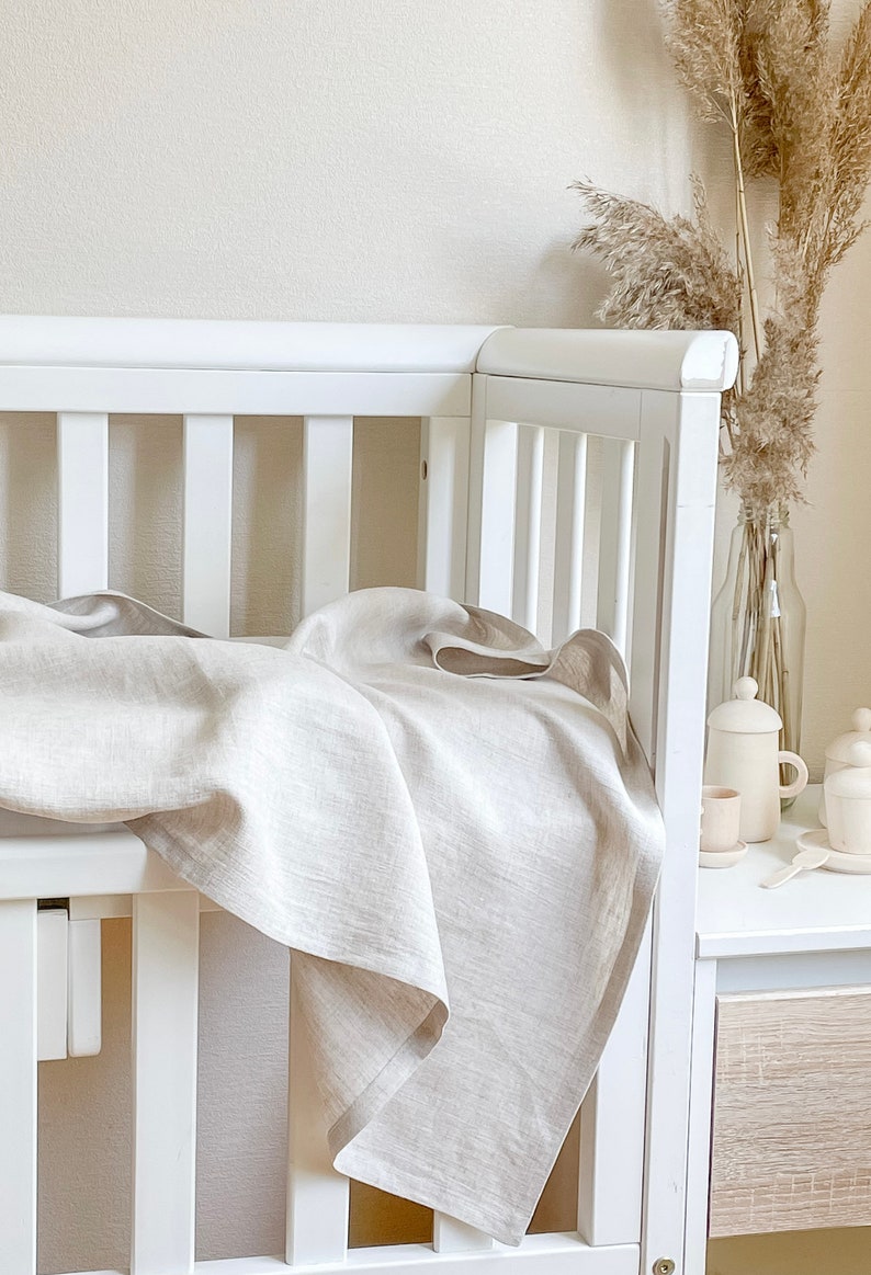 May include: A white crib with a light beige linen blanket draped over the side. The blanket has a soft, textured appearance. A vase of dried pampas grass and a small tea set are on a nearby white table.