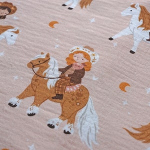 May include: A pink fabric with a repeating pattern of girls riding horses. The girls are wearing cowboy hats and dresses. The horses are white and brown with white manes and tails. The background is a light pink with small white stars and crescent moons.
