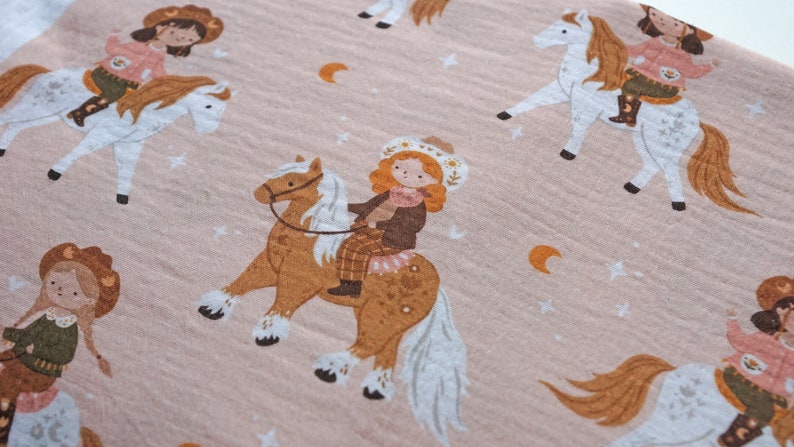 May include: A pink fabric with a repeating pattern of girls riding horses. The girls are wearing cowboy hats and dresses. The horses are white and brown with white manes and tails. The background is a light pink with small white stars and crescent moons.