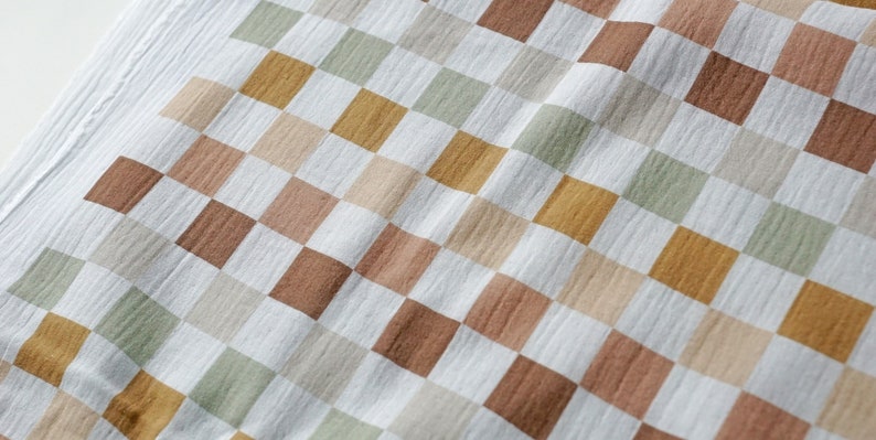 Op de afbeelding: Een witte stof met een geruit patroon in tinten bruin, beige en groen. De stof heeft een textuur, licht gerimpeld uiterlijk.