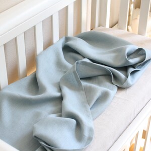 May include: A light blue linen blanket draped over a crib mattress. The blanket is soft and flowing, with visible texture. The crib is white, with a slatted design. The overall aesthetic is simple and calming, suitable for a nursery.