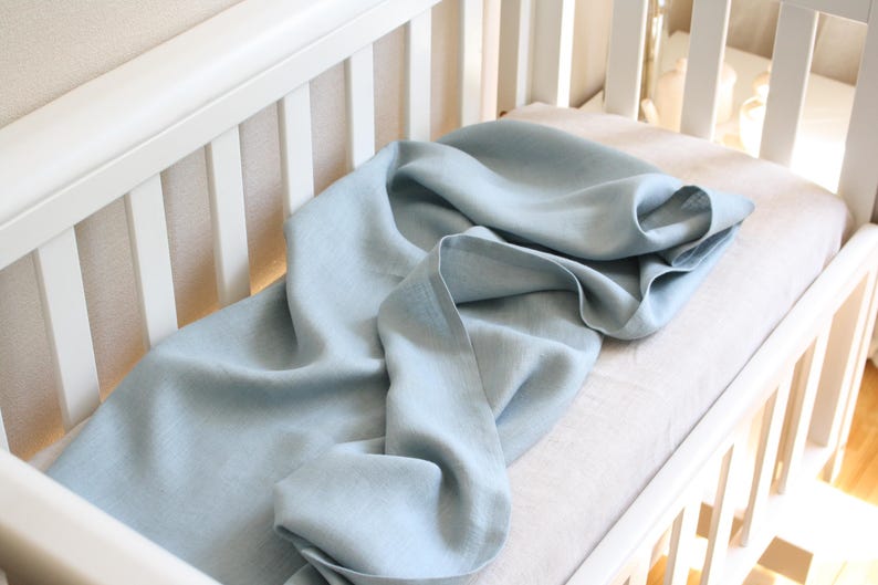 May include: A light blue linen blanket draped over a crib mattress. The blanket is soft and flowing, with visible texture. The crib is white, with a slatted design. The overall aesthetic is simple and calming, suitable for a nursery.