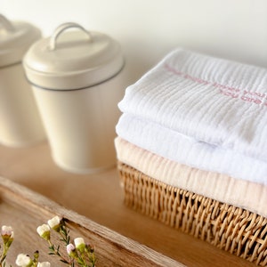 May include: A white muslin swaddle blanket with pink embroidery that reads "You are so loved" is folded and placed in a wicker basket. The basket is on a wooden tray with a sprig of white flowers.