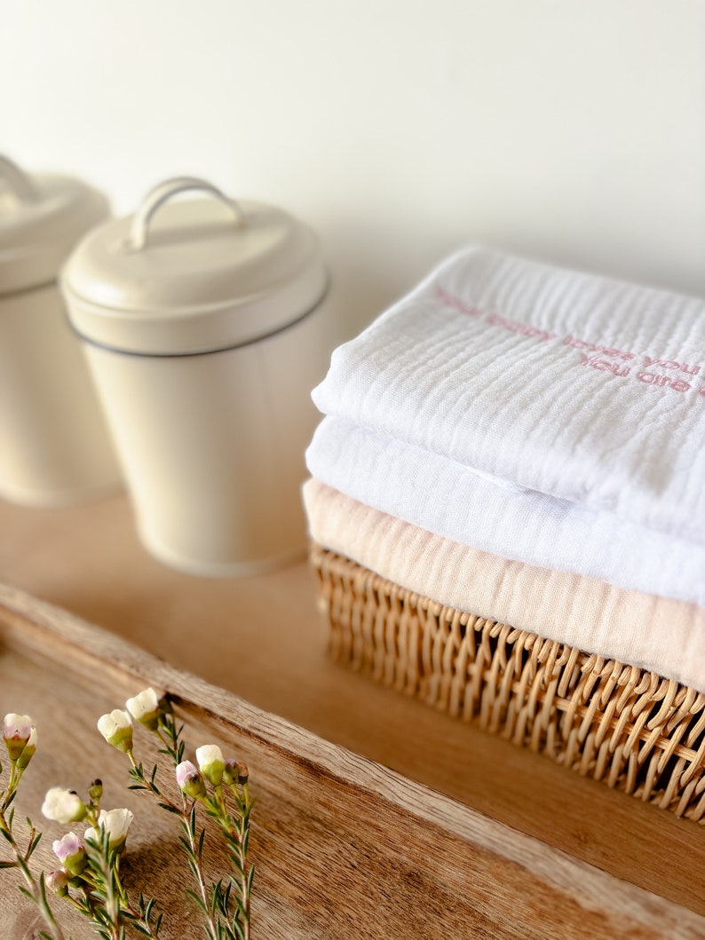 May include: A white muslin swaddle blanket with pink embroidery that reads "You are so loved" is folded and placed in a wicker basket. The basket is on a wooden tray with a sprig of white flowers.