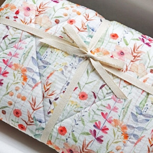 Peut inclure: Une courtepointe bébé en mousseline blanche avec un imprimé floral dans les tons rose, orange et vert. La courtepointe est nouée avec un ruban crème.