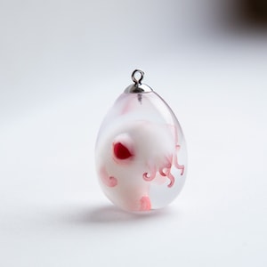 Puede incluir: Un colgante de vidrio transparente en forma de huevo que contiene una criatura blanca y rosa. La criatura tiene un corazón rojo y tentáculos rosas.