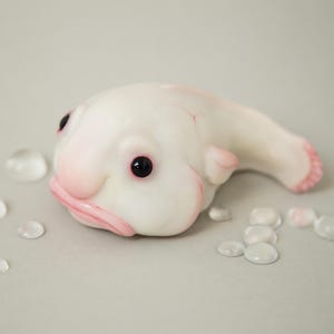 May include: A white, hand-painted sculpture of a blobfish with black eyes and a pink mouth. The sculpture is lying on its side on a light gray surface.