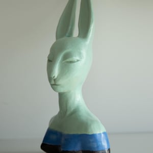 Op de afbeelding: Een keramische sculptuur van een mensachtige figuur met lange konijnenoren. De figuur is geschilderd in tinten groen, blauw en zwart. De sculptuur heeft een serene uitdrukking met gesloten ogen en een lange nek. De buste is een uniek kunstwerk.