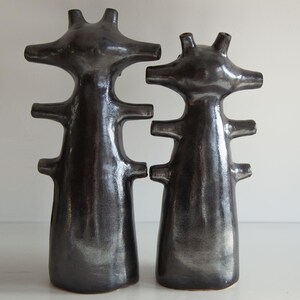 Op de afbeelding: Twee donkergrijze keramische sculpturen met een abstract ontwerp. Elke sculptuur heeft een slank lichaam met horizontale uitsteeksels en een kopachtig bovenstuk met drie punten. De sculpturen staan op een wit oppervlak.